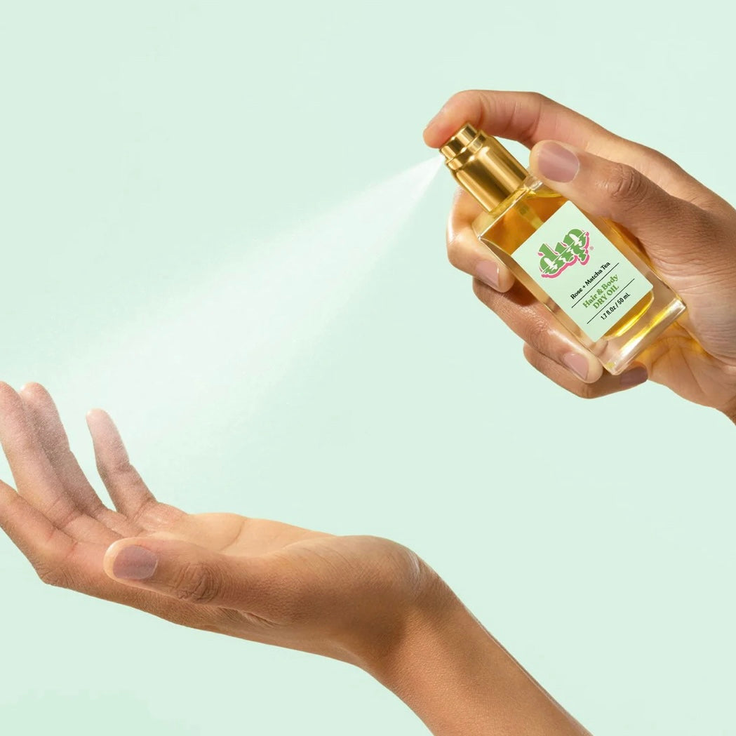 hand holding glass bottle of hair and body dry oil spraying mist over other hand on green background. 